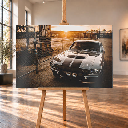 Silbergraues klassisches Fastback mit schwarzen Längsstreifen, schräg von vorn fotografiert, vor Maschendrahtzaun und Tor auf einem Industriegelände bei tief stehender, warm leuchtender Sonne; Visualisierung als Alu gebürstet-Wandbild, auf einer Holzstaffelei in einem hellen Galerieraum.