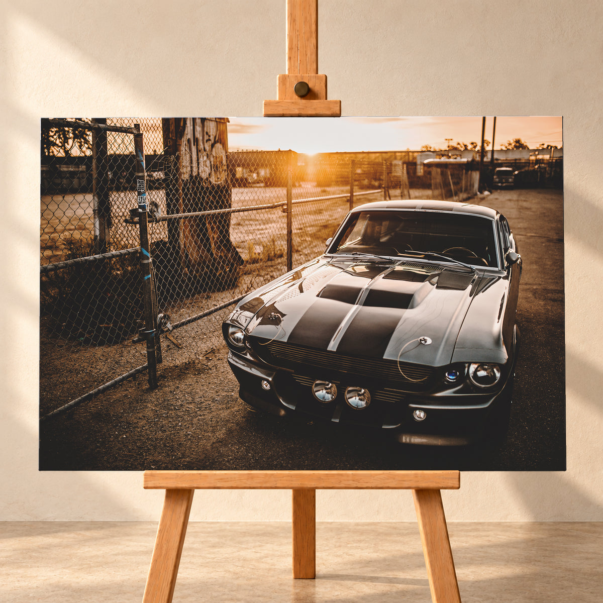 Silbergraues klassisches Fastback mit schwarzen Längsstreifen, schräg von vorn fotografiert, vor Maschendrahtzaun und Tor auf einem Industriegelände bei tief stehender, warm leuchtender Sonne; Visualisierung als Leinwand-Wandbild, frontal auf einer Holzstaffelei.