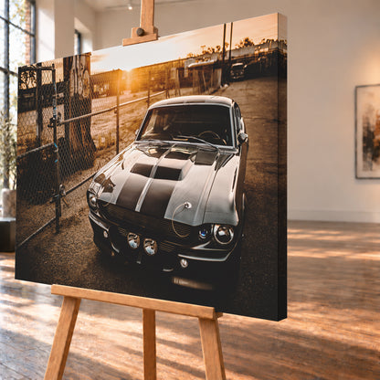 Silbergraues klassisches Fastback mit schwarzen Längsstreifen, schräg von vorn fotografiert, vor Maschendrahtzaun und Tor auf einem Industriegelände bei tief stehender, warm leuchtender Sonne; Visualisierung als Leinwand-Wandbild, schräg von der Seite auf einer Holzstaffelei.