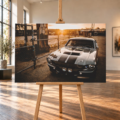 Silbergraues klassisches Fastback mit schwarzen Längsstreifen, schräg von vorn fotografiert, vor Maschendrahtzaun und Tor auf einem Industriegelände bei tief stehender, warm leuchtender Sonne; Visualisierung als Leinwand-Wandbild, auf einer Holzstaffelei in einem hellen Galerieraum.