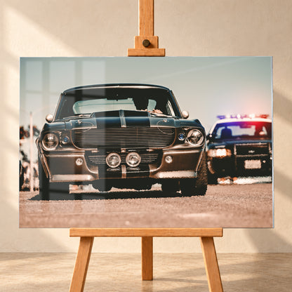 Dunkler Eleanor Ford Mustang Shelby GT500 in tiefer Frontalansicht auf heller Fahrbahn, mit dunklen Längsstreifen, Motorhaubenaufsatz, Rundscheinwerfern und zwei Zusatzleuchten. Rechts dahinter steht unscharf ein dunkles Fahrzeug mit rot-blauer Lichtleiste; Visualisierung als Acrylglas-Wandbild, frontal auf einer Holzstaffelei.