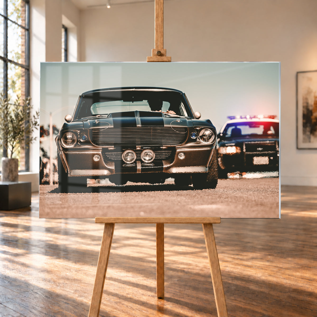 Dunkler Eleanor Ford Mustang Shelby GT500 in tiefer Frontalansicht auf heller Fahrbahn, mit dunklen Längsstreifen, Motorhaubenaufsatz, Rundscheinwerfern und zwei Zusatzleuchten. Rechts dahinter steht unscharf ein dunkles Fahrzeug mit rot-blauer Lichtleiste; Visualisierung als Acrylglas-Wandbild, auf einer Holzstaffelei in einem hellen Galerieraum.