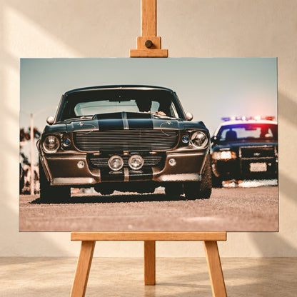 Dunkler Eleanor Ford Mustang Shelby GT500 in tiefer Frontalansicht auf heller Fahrbahn, mit dunklen Längsstreifen, Motorhaubenaufsatz, Rundscheinwerfern und zwei Zusatzleuchten. Rechts dahinter steht unscharf ein dunkles Fahrzeug mit rot-blauer Lichtleiste; Visualisierung als Leinwand-Wandbild, frontal auf einer Holzstaffelei.