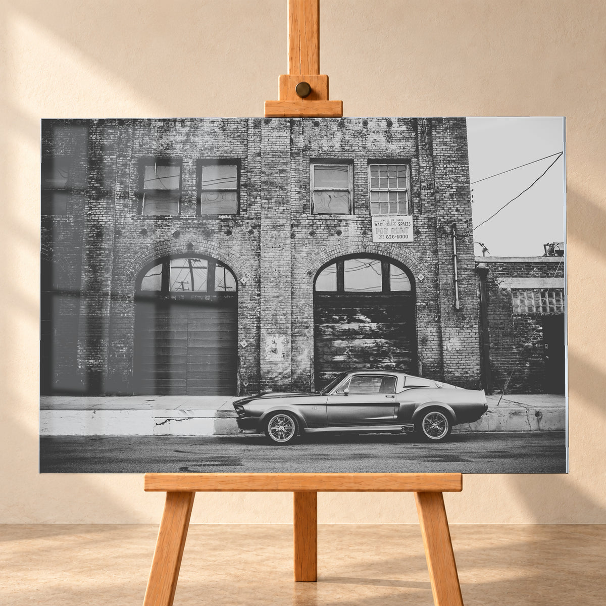 Schwarz-Weiß-Aufnahme von Eleanor in vollständiger Seitenansicht vor einer verwitterten Backsteinfassade mit großen Toren und Fenstern an einer leeren Straße; Visualisierung als Acrylglas-Wandbild, frontal auf einer Holzstaffelei.