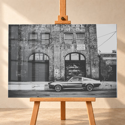 Schwarz-Weiß-Aufnahme von Eleanor in vollständiger Seitenansicht vor einer verwitterten Backsteinfassade mit großen Toren und Fenstern an einer leeren Straße; Visualisierung als Acrylglas-Wandbild, frontal auf einer Holzstaffelei.
