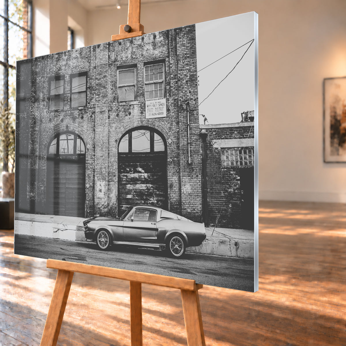 Schwarz-Weiß-Aufnahme von Eleanor in vollständiger Seitenansicht vor einer verwitterten Backsteinfassade mit großen Toren und Fenstern an einer leeren Straße; Visualisierung als Acrylglas-Wandbild, schräg von der Seite auf einer Holzstaffelei.
