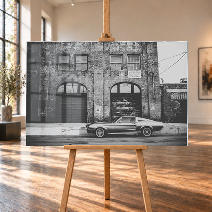 Schwarz-Weiß-Aufnahme von Eleanor in vollständiger Seitenansicht vor einer verwitterten Backsteinfassade mit großen Toren und Fenstern an einer leeren Straße; Visualisierung als Acrylglas-Wandbild, auf einer Holzstaffelei in einem hellen Galerieraum.