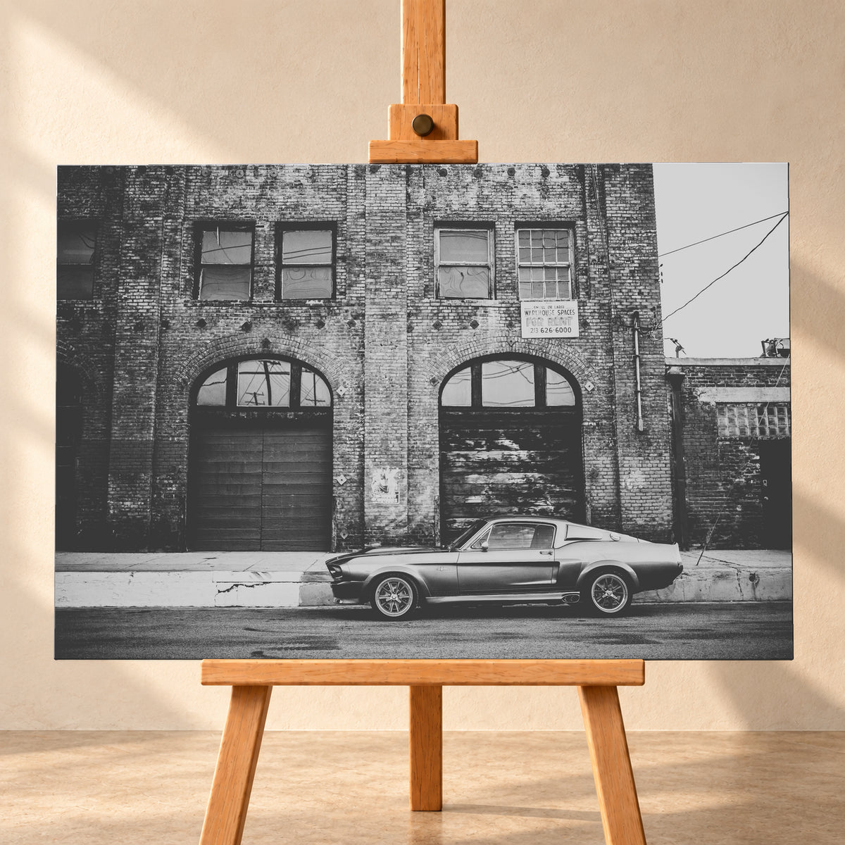 Schwarz-Weiß-Aufnahme von Eleanor in vollständiger Seitenansicht vor einer verwitterten Backsteinfassade mit großen Toren und Fenstern an einer leeren Straße; Visualisierung als Leinwand-Wandbild, frontal auf einer Holzstaffelei.
