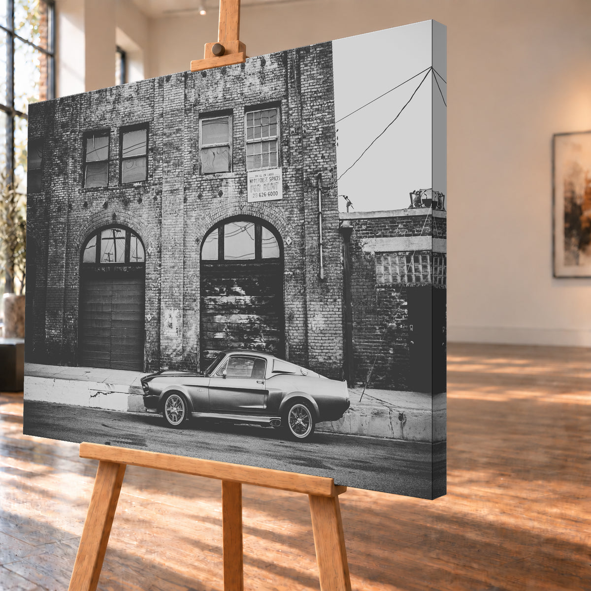 Schwarz-Weiß-Aufnahme von Eleanor in vollständiger Seitenansicht vor einer verwitterten Backsteinfassade mit großen Toren und Fenstern an einer leeren Straße; Visualisierung als Leinwand-Wandbild, schräg von der Seite auf einer Holzstaffelei.