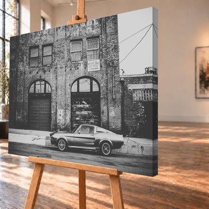 Schwarz-Weiß-Aufnahme von Eleanor in vollständiger Seitenansicht vor einer verwitterten Backsteinfassade mit großen Toren und Fenstern an einer leeren Straße; Visualisierung als Leinwand-Wandbild, schräg von der Seite auf einer Holzstaffelei.