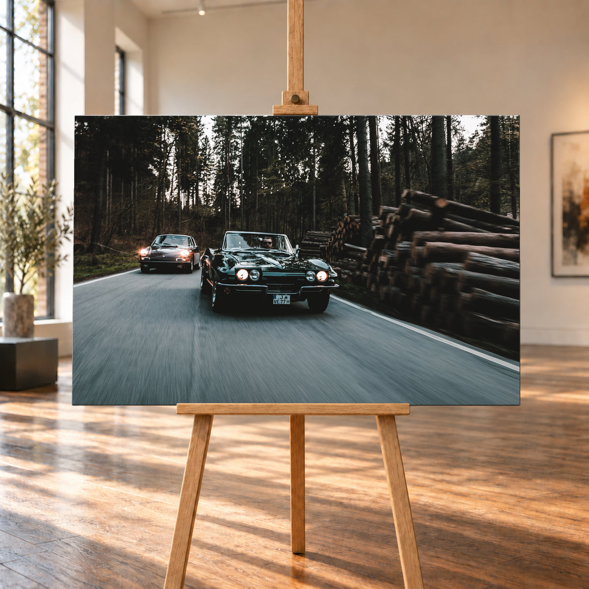 Eine dunkelgrüne 1967 Chevrolet Corvette C2 fährt frontal im Vordergrund einer Waldstraße; links dahinter ist ein dunkelroter 1967 Porsche 911 S Softwindow zu sehen. Die Scheinwerfer leuchten, Fahrer sind sichtbar. Hohe Bäume, gestapelte Baumstämme und Bewegungsunschärfe prägen die Szene; Visualisierung als Leinwand-Wandbild, auf einer Holzstaffelei in einem hellen Galerieraum.