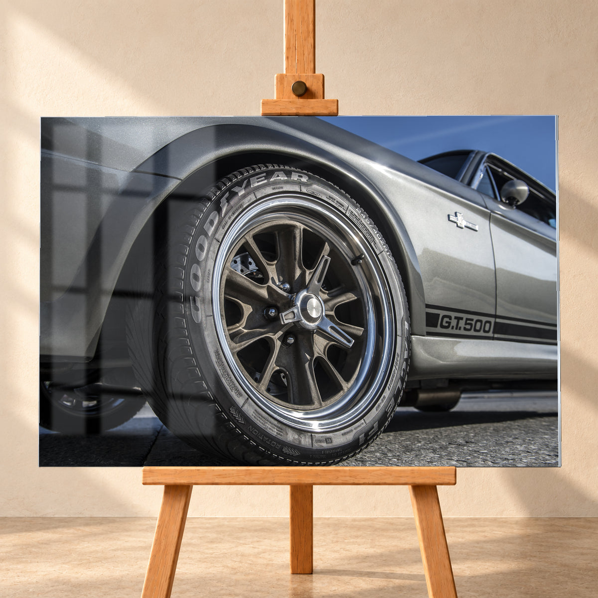 Nahaufnahme des Vorderrads eines silbergrauen Ford Mustang Shelby GT500 aus tiefer Perspektive. Dunkle Speichenfelge und Reifen dominieren das Bild; dahinter sind der Seitenstreifen mit „G.T.500“, Tür, Fenster, Außenspiegel und blauer Himmel zu sehen; Visualisierung als Acrylglas-Wandbild, frontal auf einer Holzstaffelei.