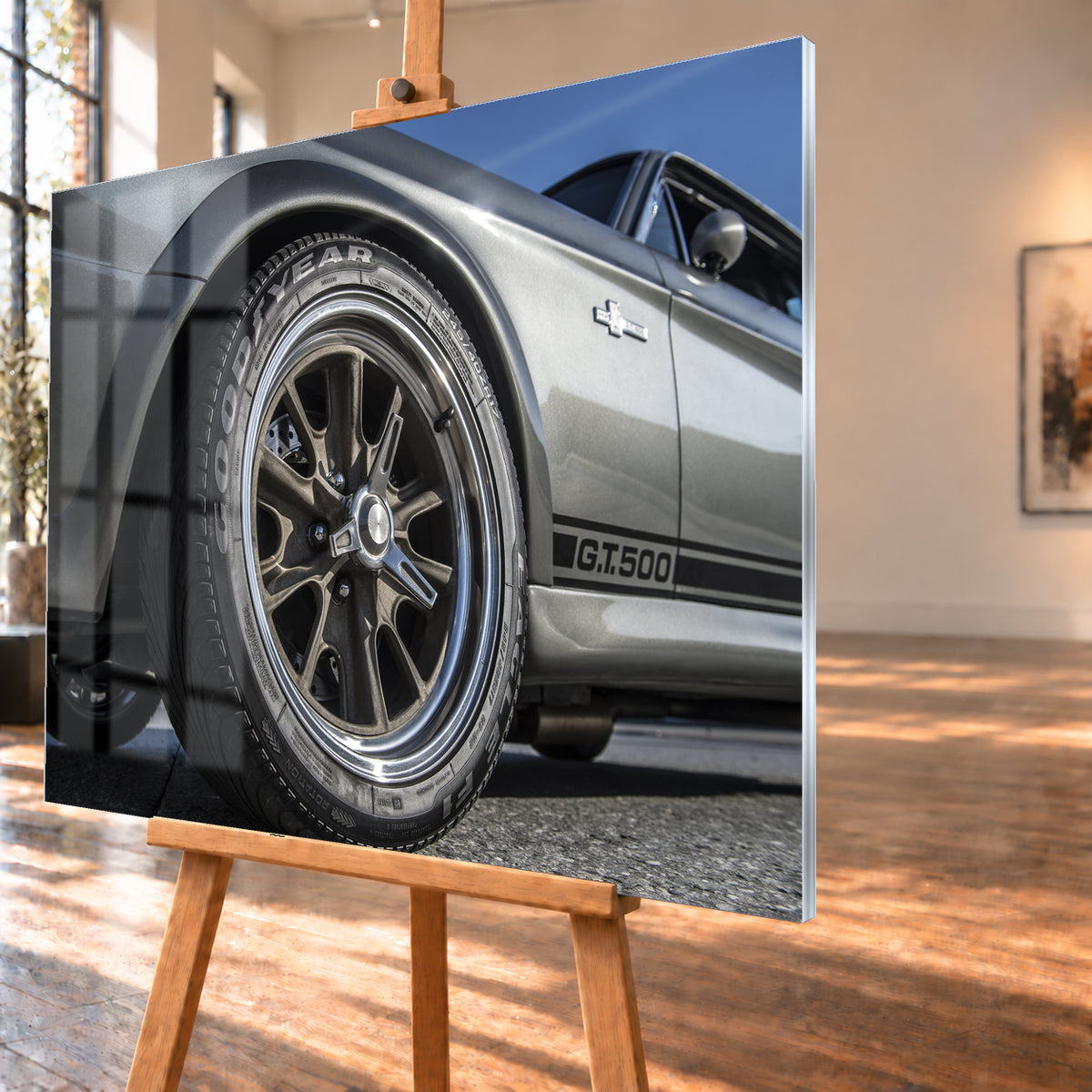 Nahaufnahme des Vorderrads eines silbergrauen Ford Mustang Shelby GT500 aus tiefer Perspektive. Dunkle Speichenfelge und Reifen dominieren das Bild; dahinter sind der Seitenstreifen mit „G.T.500“, Tür, Fenster, Außenspiegel und blauer Himmel zu sehen; Visualisierung als Acrylglas-Wandbild, schräg von der Seite auf einer Holzstaffelei.