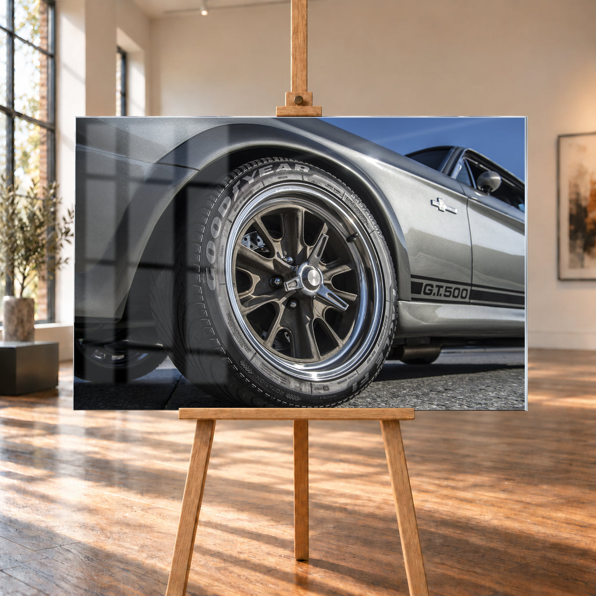 Nahaufnahme des Vorderrads eines silbergrauen Ford Mustang Shelby GT500 aus tiefer Perspektive. Dunkle Speichenfelge und Reifen dominieren das Bild; dahinter sind der Seitenstreifen mit „G.T.500“, Tür, Fenster, Außenspiegel und blauer Himmel zu sehen; Visualisierung als Acrylglas-Wandbild, auf einer Holzstaffelei in einem hellen Galerieraum.