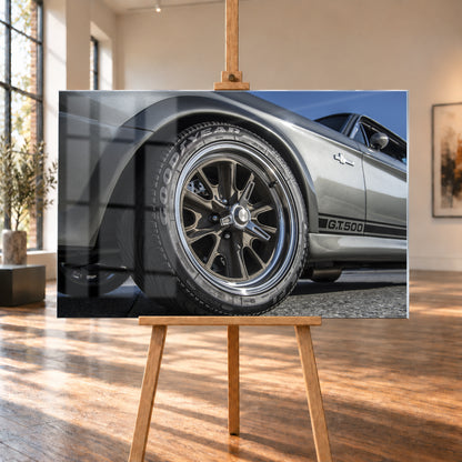 Nahaufnahme des Vorderrads eines silbergrauen Ford Mustang Shelby GT500 aus tiefer Perspektive. Dunkle Speichenfelge und Reifen dominieren das Bild; dahinter sind der Seitenstreifen mit „G.T.500“, Tür, Fenster, Außenspiegel und blauer Himmel zu sehen; Visualisierung als Acrylglas-Wandbild, auf einer Holzstaffelei in einem hellen Galerieraum.