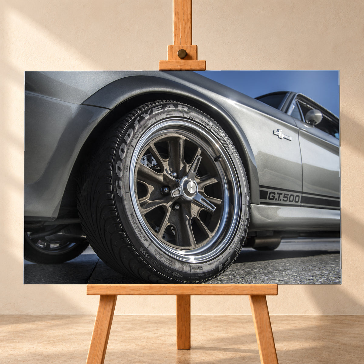 Nahaufnahme des Vorderrads eines silbergrauen Ford Mustang Shelby GT500 aus tiefer Perspektive. Dunkle Speichenfelge und Reifen dominieren das Bild; dahinter sind der Seitenstreifen mit „G.T.500“, Tür, Fenster, Außenspiegel und blauer Himmel zu sehen; Visualisierung als Alu gebürstet-Wandbild, frontal auf einer Holzstaffelei.