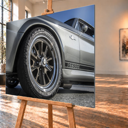 Nahaufnahme des Vorderrads eines silbergrauen Ford Mustang Shelby GT500 aus tiefer Perspektive. Dunkle Speichenfelge und Reifen dominieren das Bild; dahinter sind der Seitenstreifen mit „G.T.500“, Tür, Fenster, Außenspiegel und blauer Himmel zu sehen; Visualisierung als Alu gebürstet-Wandbild, schräg von der Seite auf einer Holzstaffelei.