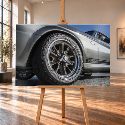 Nahaufnahme des Vorderrads eines silbergrauen Ford Mustang Shelby GT500 aus tiefer Perspektive. Dunkle Speichenfelge und Reifen dominieren das Bild; dahinter sind der Seitenstreifen mit „G.T.500“, Tür, Fenster, Außenspiegel und blauer Himmel zu sehen; Visualisierung als Alu gebürstet-Wandbild, auf einer Holzstaffelei in einem hellen Galerieraum.
