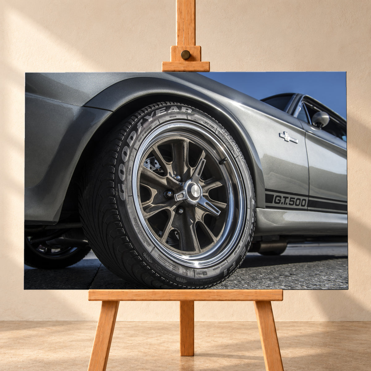 Nahaufnahme des Vorderrads eines silbergrauen Ford Mustang Shelby GT500 aus tiefer Perspektive. Dunkle Speichenfelge und Reifen dominieren das Bild; dahinter sind der Seitenstreifen mit „G.T.500“, Tür, Fenster, Außenspiegel und blauer Himmel zu sehen; Visualisierung als Leinwand-Wandbild, frontal auf einer Holzstaffelei.