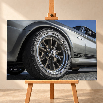 Nahaufnahme des Vorderrads eines silbergrauen Ford Mustang Shelby GT500 aus tiefer Perspektive. Dunkle Speichenfelge und Reifen dominieren das Bild; dahinter sind der Seitenstreifen mit „G.T.500“, Tür, Fenster, Außenspiegel und blauer Himmel zu sehen; Visualisierung als Leinwand-Wandbild, frontal auf einer Holzstaffelei.