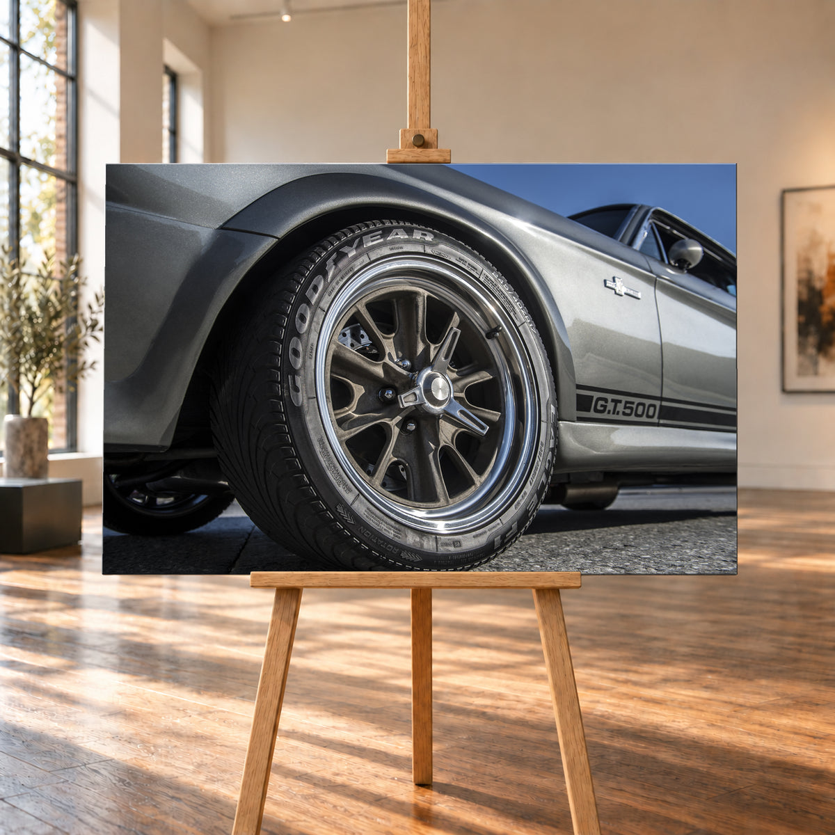 Nahaufnahme des Vorderrads eines silbergrauen Ford Mustang Shelby GT500 aus tiefer Perspektive. Dunkle Speichenfelge und Reifen dominieren das Bild; dahinter sind der Seitenstreifen mit „G.T.500“, Tür, Fenster, Außenspiegel und blauer Himmel zu sehen; Visualisierung als Leinwand-Wandbild, auf einer Holzstaffelei in einem hellen Galerieraum.