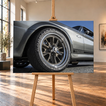 Nahaufnahme des Vorderrads eines silbergrauen Ford Mustang Shelby GT500 aus tiefer Perspektive. Dunkle Speichenfelge und Reifen dominieren das Bild; dahinter sind der Seitenstreifen mit „G.T.500“, Tür, Fenster, Außenspiegel und blauer Himmel zu sehen; Visualisierung als Leinwand-Wandbild, auf einer Holzstaffelei in einem hellen Galerieraum.