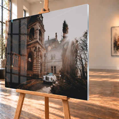 Weißes Mercedes 300 SE Cabriolet von 1965 in Front-Dreiviertelansicht auf einer feuchten Auffahrt vor einem historischen Steingebäude; tief stehende Sonne strahlt rechts durch einen hohen Baum, kahle Äste und kleine Schneereste säumen den Weg; Visualisierung als Acrylglas-Wandbild, schräg von der Seite auf einer Holzstaffelei.