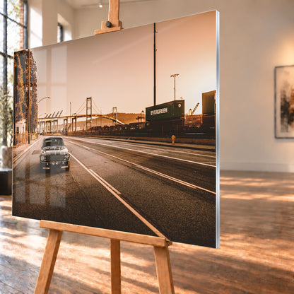 Dunkler klassischer Ford Mustang mit zwei hellen Längsstreifen frontal auf einer breiten Hafenstraße; im Hintergrund stehen eine Hängebrücke, Container und Kräne unter warmem, goldfarbenem Himmel; Visualisierung als Acrylglas-Wandbild, schräg von der Seite auf einer Holzstaffelei.