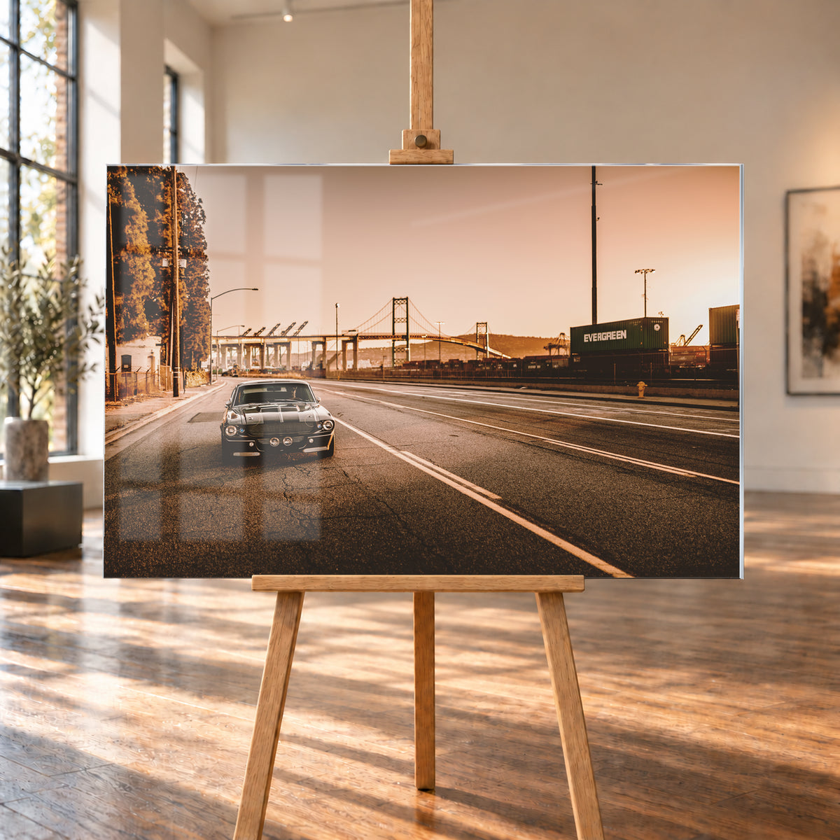 Dunkler klassischer Ford Mustang mit zwei hellen Längsstreifen frontal auf einer breiten Hafenstraße; im Hintergrund stehen eine Hängebrücke, Container und Kräne unter warmem, goldfarbenem Himmel; Visualisierung als Acrylglas-Wandbild, auf einer Holzstaffelei in einem hellen Galerieraum.