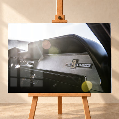 Nahaufnahme des schwarzen Armaturenbretts von Eleanor mit Shelby-, Cobra- und G.T.-500-Emblemen. Helles Gegenlicht fällt von links oben ein und erzeugt grüne, gelbe und rötliche Lichtreflexe; Visualisierung als Acrylglas-Wandbild, frontal auf einer Holzstaffelei.