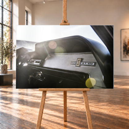 Nahaufnahme des schwarzen Armaturenbretts von Eleanor mit Shelby-, Cobra- und G.T.-500-Emblemen. Helles Gegenlicht fällt von links oben ein und erzeugt grüne, gelbe und rötliche Lichtreflexe; Visualisierung als Alu gebürstet-Wandbild, auf einer Holzstaffelei in einem hellen Galerieraum.