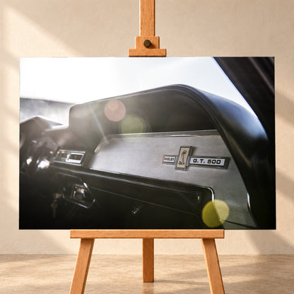 Nahaufnahme des schwarzen Armaturenbretts von Eleanor mit Shelby-, Cobra- und G.T.-500-Emblemen. Helles Gegenlicht fällt von links oben ein und erzeugt grüne, gelbe und rötliche Lichtreflexe; Visualisierung als Leinwand-Wandbild, frontal auf einer Holzstaffelei.