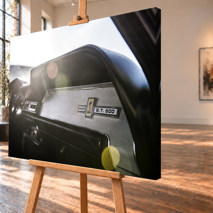 Nahaufnahme des schwarzen Armaturenbretts von Eleanor mit Shelby-, Cobra- und G.T.-500-Emblemen. Helles Gegenlicht fällt von links oben ein und erzeugt grüne, gelbe und rötliche Lichtreflexe; Visualisierung als Leinwand-Wandbild, schräg von der Seite auf einer Holzstaffelei.