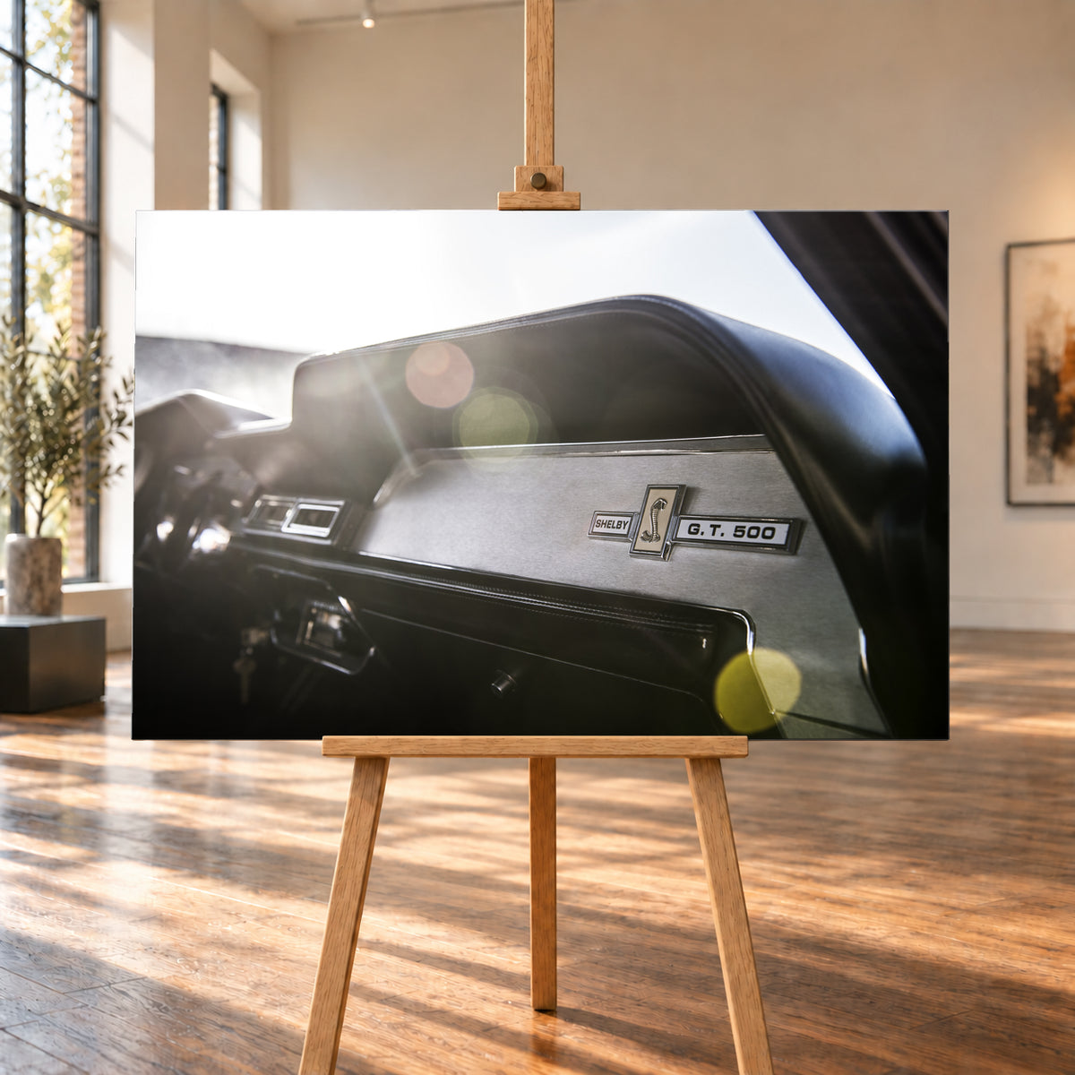 Nahaufnahme des schwarzen Armaturenbretts von Eleanor mit Shelby-, Cobra- und G.T.-500-Emblemen. Helles Gegenlicht fällt von links oben ein und erzeugt grüne, gelbe und rötliche Lichtreflexe; Visualisierung als Leinwand-Wandbild, auf einer Holzstaffelei in einem hellen Galerieraum.