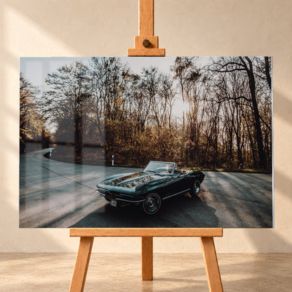 Eine dunkelgrüne 1967 Chevrolet Corvette C2 als offenes Cabriolet in Front-Dreiviertelansicht auf einer kurvigen Waldstraße. Tief stehende Sonne scheint durch kahle Bäume; Gegenlicht und Astspiegelungen liegen auf der Karosserie; Visualisierung als Acrylglas-Wandbild, frontal auf einer Holzstaffelei.