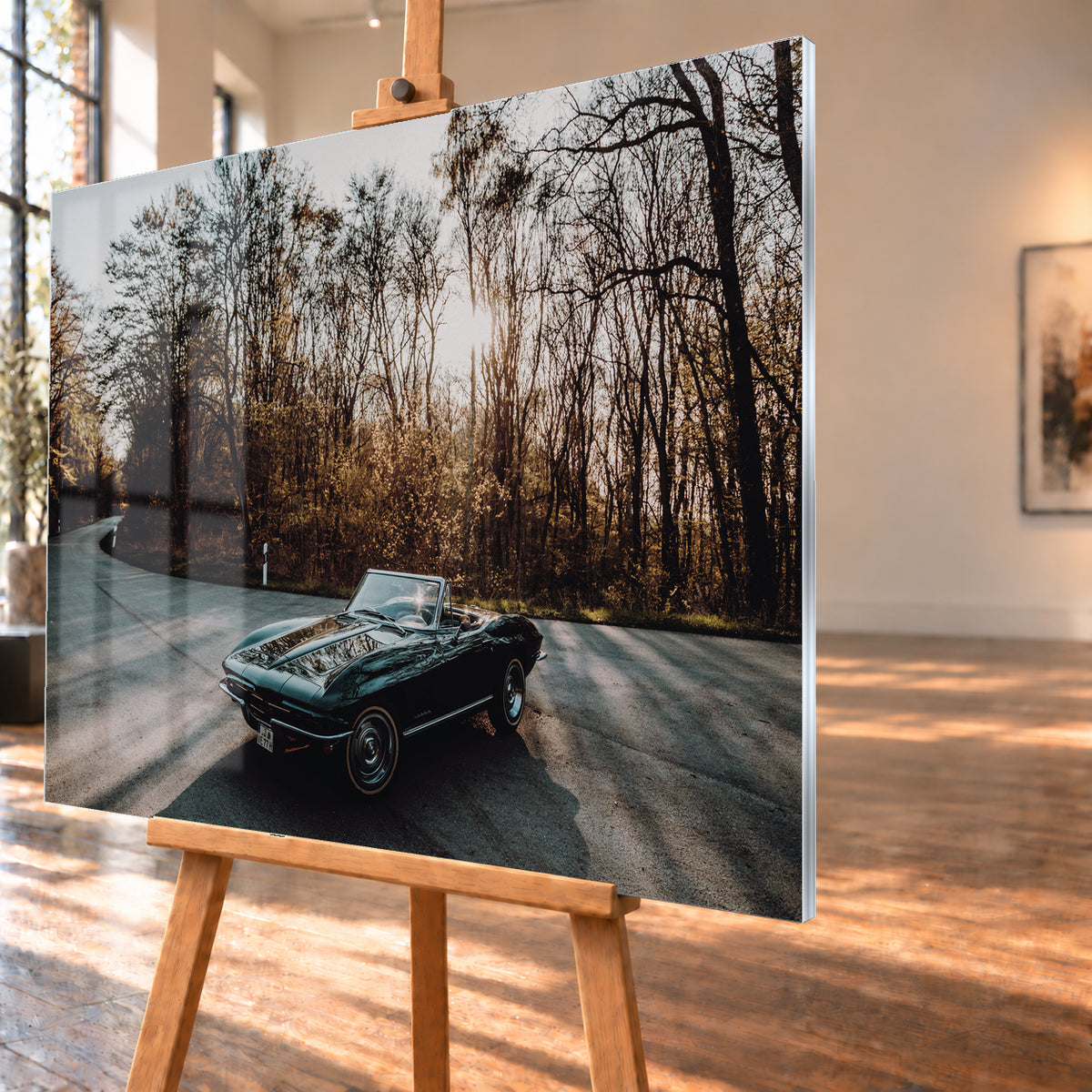 Eine dunkelgrüne 1967 Chevrolet Corvette C2 als offenes Cabriolet in Front-Dreiviertelansicht auf einer kurvigen Waldstraße. Tief stehende Sonne scheint durch kahle Bäume; Gegenlicht und Astspiegelungen liegen auf der Karosserie; Visualisierung als Acrylglas-Wandbild, schräg von der Seite auf einer Holzstaffelei.