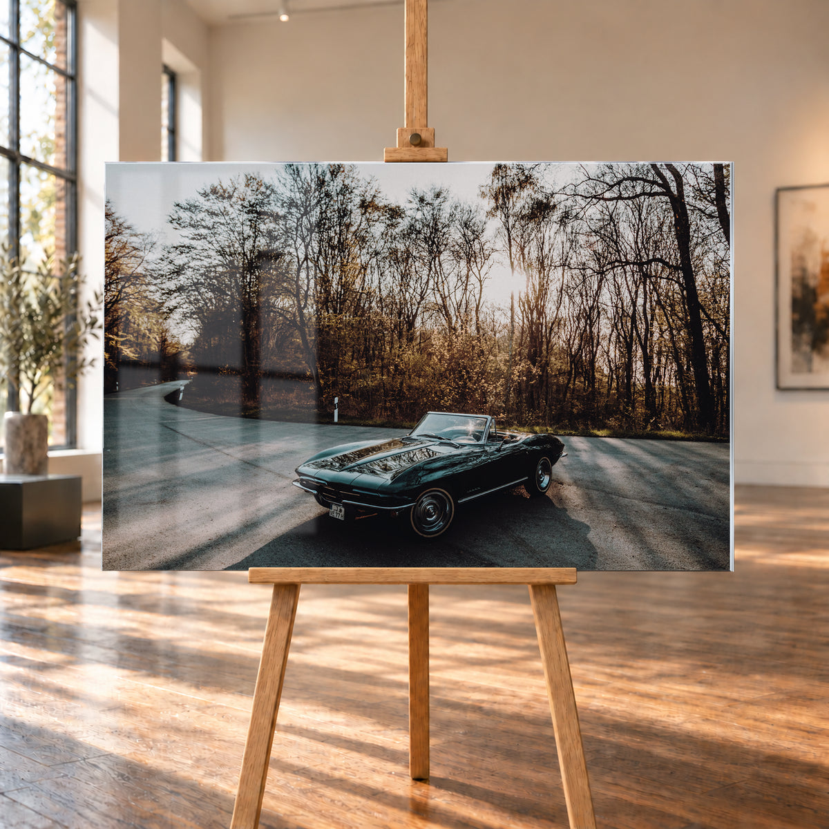 Eine dunkelgrüne 1967 Chevrolet Corvette C2 als offenes Cabriolet in Front-Dreiviertelansicht auf einer kurvigen Waldstraße. Tief stehende Sonne scheint durch kahle Bäume; Gegenlicht und Astspiegelungen liegen auf der Karosserie; Visualisierung als Acrylglas-Wandbild, auf einer Holzstaffelei in einem hellen Galerieraum.