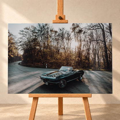 Eine dunkelgrüne 1967 Chevrolet Corvette C2 als offenes Cabriolet in Front-Dreiviertelansicht auf einer kurvigen Waldstraße. Tief stehende Sonne scheint durch kahle Bäume; Gegenlicht und Astspiegelungen liegen auf der Karosserie; Visualisierung als Alu gebürstet-Wandbild, frontal auf einer Holzstaffelei.