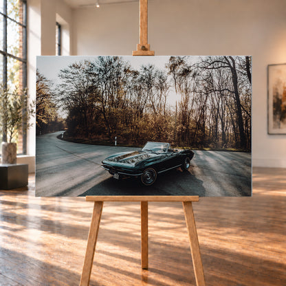 Eine dunkelgrüne 1967 Chevrolet Corvette C2 als offenes Cabriolet in Front-Dreiviertelansicht auf einer kurvigen Waldstraße. Tief stehende Sonne scheint durch kahle Bäume; Gegenlicht und Astspiegelungen liegen auf der Karosserie; Visualisierung als Alu gebürstet-Wandbild, auf einer Holzstaffelei in einem hellen Galerieraum.