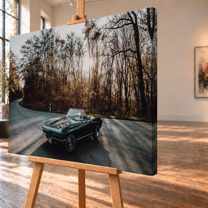Eine dunkelgrüne 1967 Chevrolet Corvette C2 als offenes Cabriolet in Front-Dreiviertelansicht auf einer kurvigen Waldstraße. Tief stehende Sonne scheint durch kahle Bäume; Gegenlicht und Astspiegelungen liegen auf der Karosserie; Visualisierung als Leinwand-Wandbild, schräg von der Seite auf einer Holzstaffelei.