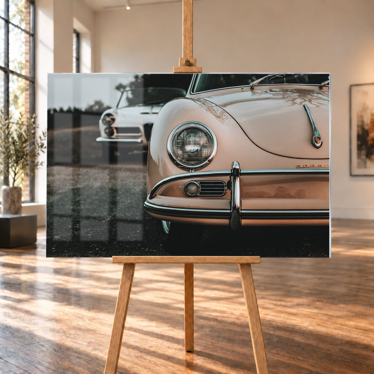 Nahaufnahme der Front eines roséfarbenen Porsche 356 A mit Rundscheinwerfer und Chromstoßfänger rechts; links dahinter steht unscharf ein heller Alfa Romeo 2000 Touring Spider auf dunklem Untergrund unter grauem Himmel; Visualisierung als Acrylglas-Wandbild, auf einer Holzstaffelei in einem hellen Galerieraum.