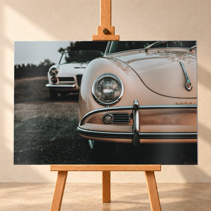 Nahaufnahme der Front eines roséfarbenen Porsche 356 A mit Rundscheinwerfer und Chromstoßfänger rechts; links dahinter steht unscharf ein heller Alfa Romeo 2000 Touring Spider auf dunklem Untergrund unter grauem Himmel; Visualisierung als Alu gebürstet-Wandbild, frontal auf einer Holzstaffelei.