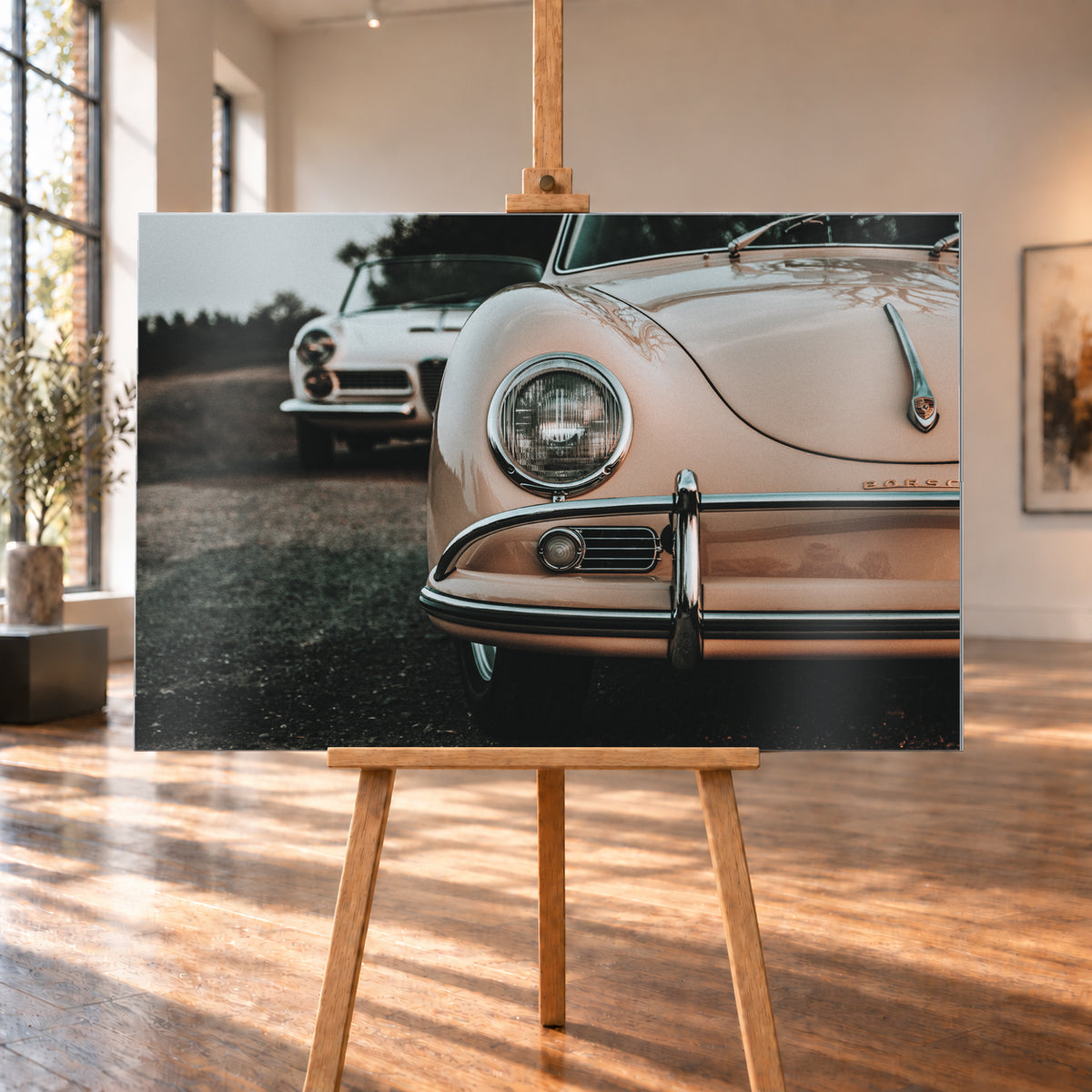 Nahaufnahme der Front eines roséfarbenen Porsche 356 A mit Rundscheinwerfer und Chromstoßfänger rechts; links dahinter steht unscharf ein heller Alfa Romeo 2000 Touring Spider auf dunklem Untergrund unter grauem Himmel; Visualisierung als Alu gebürstet-Wandbild, auf einer Holzstaffelei in einem hellen Galerieraum.