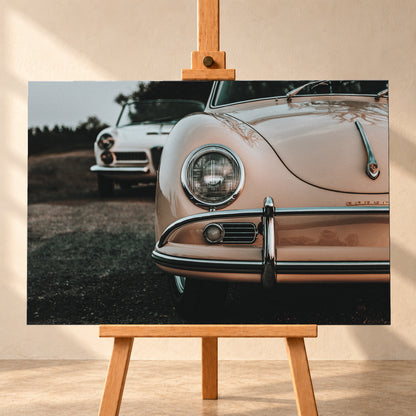 Nahaufnahme der Front eines roséfarbenen Porsche 356 A mit Rundscheinwerfer und Chromstoßfänger rechts; links dahinter steht unscharf ein heller Alfa Romeo 2000 Touring Spider auf dunklem Untergrund unter grauem Himmel; Visualisierung als Leinwand-Wandbild, frontal auf einer Holzstaffelei.