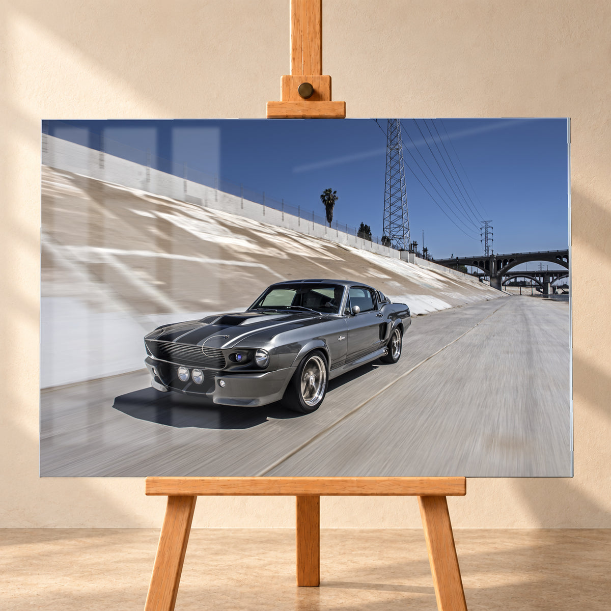 Metallisch grauer Ford Mustang Shelby GT500 mit schwarzen Doppelstreifen in niedriger Front-Dreiviertelansicht in einem hellen Betonkanal. Fahrbahn und Kanalwand sind bewegungsunscharf; Strommasten, Leitungen, Palmen und Bogenbrücken stehen unter blauem Himmel; Visualisierung als Acrylglas-Wandbild, frontal auf einer Holzstaffelei.