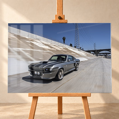 Metallisch grauer Ford Mustang Shelby GT500 mit schwarzen Doppelstreifen in niedriger Front-Dreiviertelansicht in einem hellen Betonkanal. Fahrbahn und Kanalwand sind bewegungsunscharf; Strommasten, Leitungen, Palmen und Bogenbrücken stehen unter blauem Himmel; Visualisierung als Acrylglas-Wandbild, frontal auf einer Holzstaffelei.
