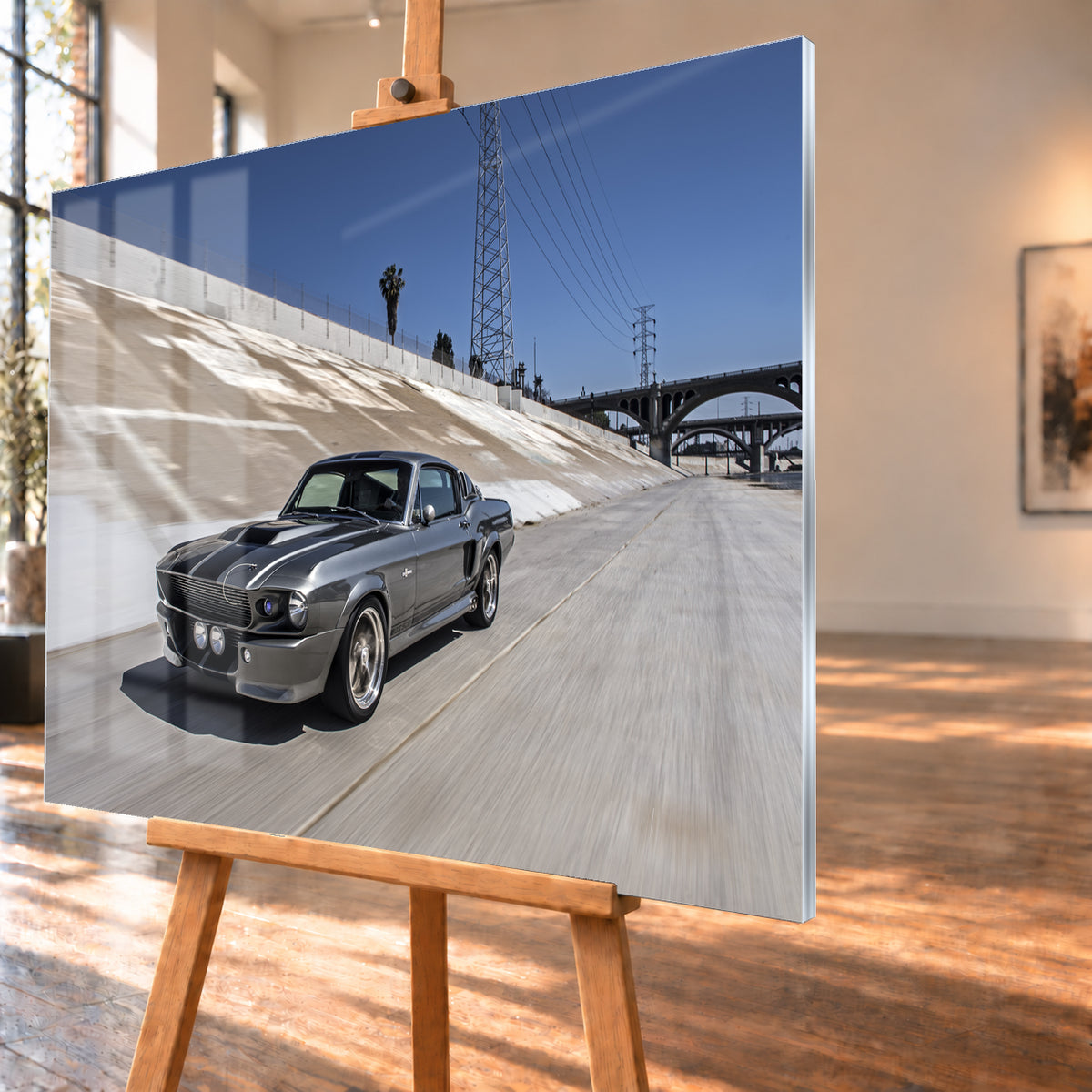Metallisch grauer Ford Mustang Shelby GT500 mit schwarzen Doppelstreifen in niedriger Front-Dreiviertelansicht in einem hellen Betonkanal. Fahrbahn und Kanalwand sind bewegungsunscharf; Strommasten, Leitungen, Palmen und Bogenbrücken stehen unter blauem Himmel; Visualisierung als Acrylglas-Wandbild, schräg von der Seite auf einer Holzstaffelei.