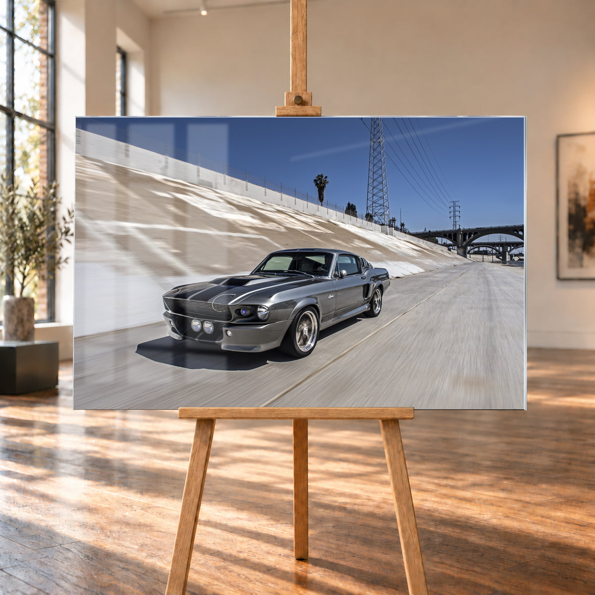 Metallisch grauer Ford Mustang Shelby GT500 mit schwarzen Doppelstreifen in niedriger Front-Dreiviertelansicht in einem hellen Betonkanal. Fahrbahn und Kanalwand sind bewegungsunscharf; Strommasten, Leitungen, Palmen und Bogenbrücken stehen unter blauem Himmel; Visualisierung als Acrylglas-Wandbild, auf einer Holzstaffelei in einem hellen Galerieraum.