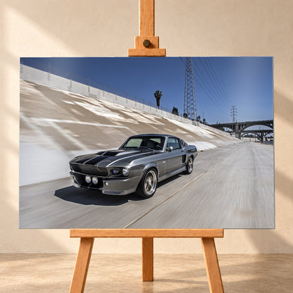 Metallisch grauer Ford Mustang Shelby GT500 mit schwarzen Doppelstreifen in niedriger Front-Dreiviertelansicht in einem hellen Betonkanal. Fahrbahn und Kanalwand sind bewegungsunscharf; Strommasten, Leitungen, Palmen und Bogenbrücken stehen unter blauem Himmel; Visualisierung als Alu gebürstet-Wandbild, frontal auf einer Holzstaffelei.