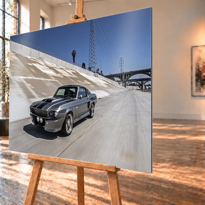Metallisch grauer Ford Mustang Shelby GT500 mit schwarzen Doppelstreifen in niedriger Front-Dreiviertelansicht in einem hellen Betonkanal. Fahrbahn und Kanalwand sind bewegungsunscharf; Strommasten, Leitungen, Palmen und Bogenbrücken stehen unter blauem Himmel; Visualisierung als Alu gebürstet-Wandbild, schräg von der Seite auf einer Holzstaffelei.